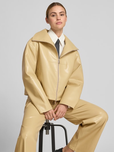 Frauenmodel trägt Jacke mit Stehkragen von Nanushka in Leder-Optik, Regular-Fit, Zweiwege-Reißverschluss, Eingrifftaschen, beige, Nahaufnahme Oberkörper. Rückenlänge 55 cm, Brustweite 55 cm, Ärmellänge 66 cm bei Größe S.