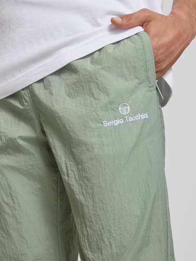 Männermodel trägt Straight Fit Stoffhose mit Logo-Stitching Modell 'Nayla' von SERGIO TACCHINI in Grün, Nahaufnahme Oberkörper. Herren Stoffhose, elastischer Bund mit Tunnelzug, elastische Beinabschlüsse, Innenbeinlänge bei Größe M 77 cm.