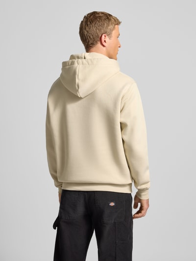 Męski model nosi Hoodie mit Label-Stitching Modell 'Camo Sleeve' od Alpha Industries w kolorze kremowym, bawełniany mix, widok całej sylwetki z tyłu. Produkt posiada regular fit, kieszeń kangurkę, długość tyłu 67 cm, rękawy 66 cm.