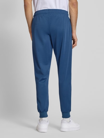Męski model nosi spodnie dresowe z mieszanki bawełny Ellesse Sweatpants mit elastischem Bund Modell 'BERTONI', krój regular fit, detal z logo, długość wewnętrzna nogawki w rozmiarze M: 76 cm, kolor niebieski, gładka tekstura, pełny widok sylwetki z tyłu.