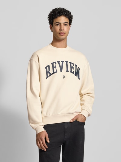 Männermodel trägt Rundhalssweat mit Varsiety Embleme von Review in Creme mit schwarzem Schriftzug, Baumwoll-Mix, lockerer Schnitt, Nahaufnahme Oberkörper.
