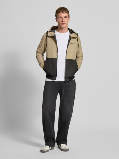 Männermodel trägt beige-schwarze Jacke mit Capuchon en ritssluiting Model 'RUSH' van JACK & JONES met logoprint en steekzakken, regular fit, ruglengte 68 cm, schouderbreedte 46 cm, borstwijdte 55 cm, mouwlengte 68 cm, ganzkörperansicht van voren.