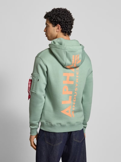 Männermodel trägt Hoodie mit Känguru-Tasche von Alpha Industries in Hellgrün, Baumwoll-Mix, Label-Patch, Regular-Fit, Ganzkörperansicht Rückansicht.