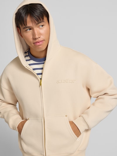 Männermodel trägt Sweatjacke mit Kapuze von ANOTHER COTTON LAB in Creme, lockerer Schnitt, Känguru-Tasche, Nahaufnahme Oberkörper. Elastischer Baumwoll-Mix mit gerippten Abschlüssen, Label-Detail, Zwei-Wege-Reißverschluss.