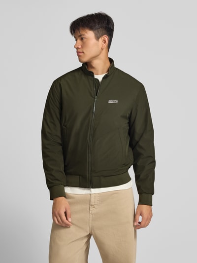 Männermodel trägt Bomberjacke mit Logo Modell 'BROOKLYN' von JACK & JONES in Oliv, Reißverschluss, glatte Struktur, Ganzkörperansicht von vorne.