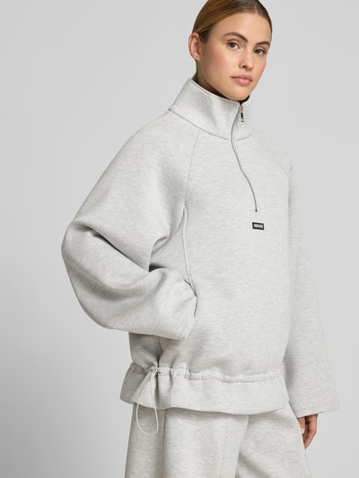 Frauenmodel trägt Sweatshirt aus Viskose-Mix von ROTATE in hellem Grau mit Stehkragen, Reißverschluss, Logo-Patch, Taillierte Passform, Detailaufnahme Oberkörper.