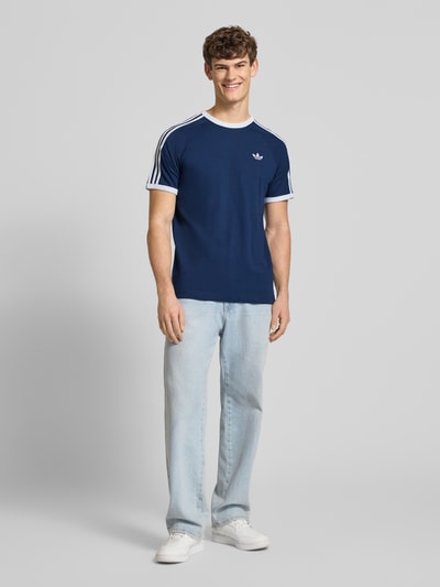 Männermodel trägt T-Shirt mit Label-Stitching von adidas Originals aus reiner Baumwolle, Regular-Fit, Gerippte-Abschlüsse, Rundhalsausschnitt, Rückenlänge 73 cm bei Größe M, dunkelblau mit weißen Details, Ganzkörperansicht von vorne.