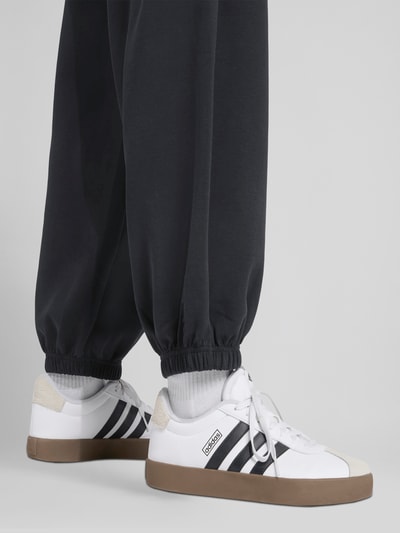 Männermodel trägt Comfort Fit Sweatpants mit Logo-Print von adidas Sportswear in Schwarz, Sweatstoff, elastischer Bund, Nahaufnahme Beine, seitliche Eingrifftaschen, Ganzkörperansicht Rückansicht.