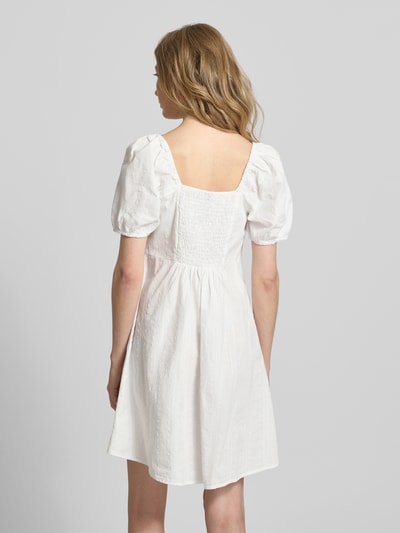 Frauenmodel trägt A-Linien Umstandskleid aus reiner Baumwolle Modell 'JUTTY' von Vero Moda, Optik: Unifarben, Struktur-Muster, Mini-Länge, Ballonärmel, elastischer Ärmelabschluss, Bundfalte, Gummizug, ganz in Weiß, Ganzkörperansicht Rückansicht.