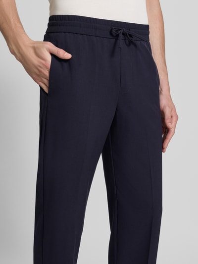 Męski model nosi Relaxed Fit Sweatpants mit Viskose-Anteil von Lindbergh, elastyczna mieszanka materiałów z dodatkiem wiskozy, kanty, spodnie w kolorze granatowym, szerokość w rozmiarze M: 41 cm, długość wewnętrzna nogawki w rozmiarze M: 79 cm, z tunelem w pasie, zbliżenie na przednią część spodni, detaliczny widok górnej części.