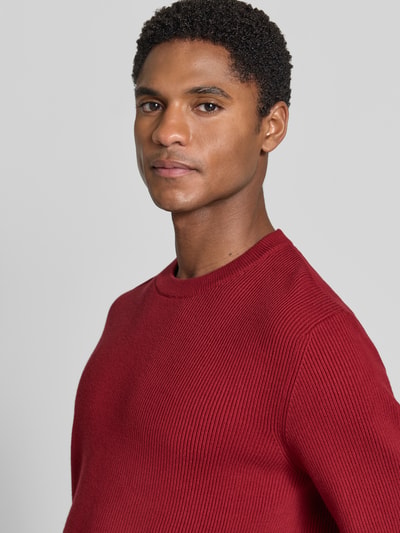 Męski model nosi Slim Fit Strickpullover Baumwoll-Mix od Lindbergh w kolorze czerwonym, efekt prążkowania, okrągły dekolt, mieszanka bawełny, długość tyłu 70 cm, długość rękawów 72 cm, zbliżenie na górną część ciała.