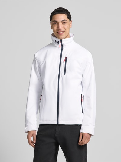 Männermodel trägt weiße Midlayer Jacke mit Stehkragen Modell 'CREW' von Helly Hansen, Gerader-Schnitt, Logo-Print, Reißverschluss, Label-Patch, Brusttasche, seitliche Reißverschlusstaschen, Ganzkörperansicht von vorne.