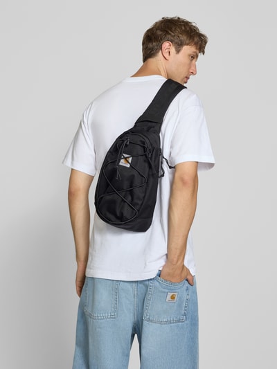 Männermodel trägt zwarte Sling Bag met logopatch Model 'Kyle' van Carhartt Work In Progress, Tunnelkoord, Hoofdvak met ritssluiting, verstelbare schouderband, Breedte 20 cm, Lengte 35 cm, Diepte 10 cm, bandlengte 90 cm, Ganzkörperansicht Rückansicht.
