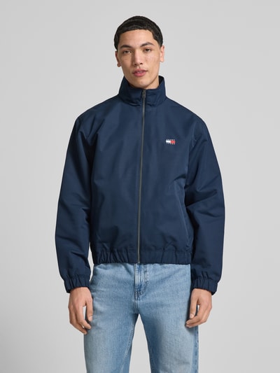 Męski model nosi Regular Fit Blouson mit Stehkragen von Tommy Jeans, efekt: jednolity kolor, krój regular fit, stójka, zapięcie: zamek błyskawiczny, naszywka z logo, długi rękaw, elastyczne zakończenia, boczne wpuszczane kieszenie, kieszeń wewnętrzna, granatowy, gładka tekstura, Ganzkörperansicht von vorne.