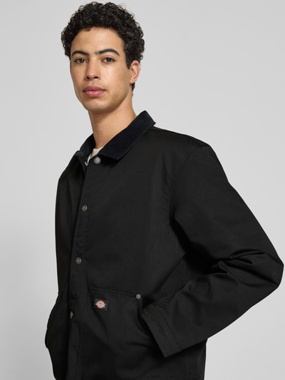Männermodel trägt Jacke mit Umlegekragen und Label-Detail von Dickies in Schwarz, Material-Mix mit Baumwoll-Anteil, Regular-Fit, Eingrifftaschen, Knopfleiste, Detailaufnahme Oberkörper. Rückenlänge bei Größe M: 75 cm, Schulterbreite: 47 cm, Brustweite: 64 cm, Ärmellänge: 66 cm.