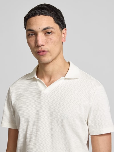Męski model nosi Regular Fit Polo-Shirt mit Strukturmuster Modell 'Heskild' von Blend z bawełny, kołnierzyk polo, krótki rękaw, fakturowany wzór, długość tyłu w rozmiarze M: 69 cm, kolor jasny, faktura lekko pofalowana, zbliżenie górnej części ciała.