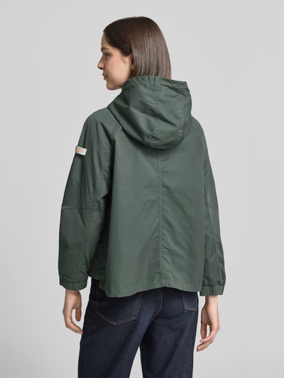 Frauenmodel trägt kurtka Jacke mit Zweiwege-Reißverschluss Modell 'DOLORES NEO' von Frieda & Freddies z mieszanka bawełny, prosty krój, kolor zielony, naszywka z logo, kaptur, kieszenie z patką, elastyczne zakończenia rękawów, regulowany dół z troczkami, długość tyłu w rozmiarze 36: 59 cm, szerokość ramion w rozmiarze 36: 48 cm, szerokość w rozmiarze 36: 50 cm, długość rękawów w rozmiarze 36: 52 cm. Ganzkörperansicht Rückansicht.