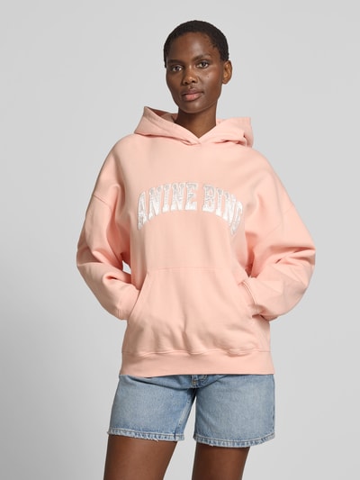 Frauenmodel trägt Oversized Hoodie mit Logo-Detail von Anine Bing aus Reine Baumwolle in Hellrosa mit Känguru-Tasche und elastischen Abschlüssen, Oversized-Fit, Rückenlänge bei Größe S 64 cm, Ganzkörperansicht von vorne.