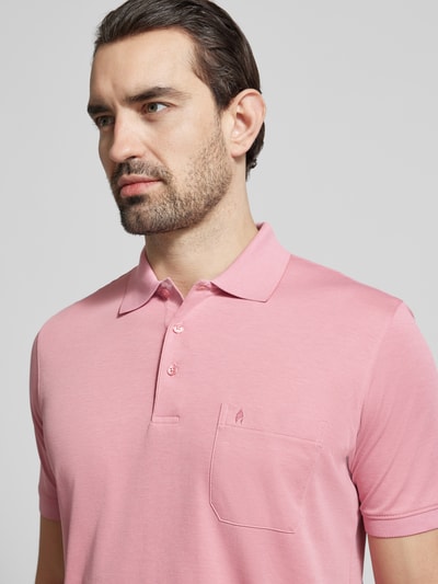 Męski model nosi Relaxed Fit Poloshirt aus Baumwoll-Mix von RAGMAN, mieszanka bawełny, kołnierzyk polo, Brustasza z wyhaftowanym logo, krótki rękaw, elastyczne zakończenia rękawów, długość tyłu w rozmiarze M 70 cm, jasnoróżowy kolor, gładka tekstura. Zbliżenie na górną część tułowia.