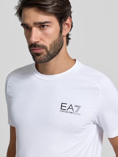 Męski t-shirt T-Shirt mit Label-Print od EA7 Emporio Armani, mieszanka materiałów z dodatkiem streczu, krój slim fit, prążkowany okrągły dekolt, nadruk z logo na piersi, długość tyłu 69 cm w rozmiarze M, zbliżenie na górną część przodu.