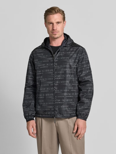 Männermodel trägt Jacke mit Logo-Muster und Zweiwege-Reißverschluss von Emporio Armani, Regular-Fit, Kapuze mit Tunnelzug, Reißverschlusstaschen, schwarz-graue Farben, glatte Textur, Rückenlänge bei Größe 50: 70 cm, Schulterbreite: 58 cm, Brustweite: 62 cm, Ärmellänge: 66 cm, Ganzkörperansicht von vorne.