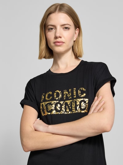 Frauenmodel trägt T-Shirt mit Rundhalsausschnitt und Print Modell 'MARICA' von Soyaconcept, Schwarz mit goldfarbenem Print, Lyocell-Elasthan-Mix, Relaxed Fit, Ärmel mit Aufschlag, Nahaufnahme Oberkörper.