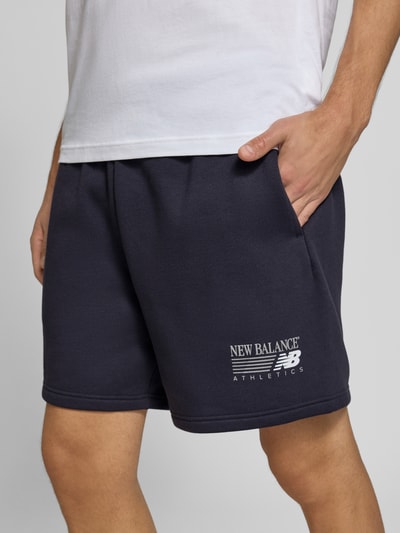 Männermodel trägt Sweatshorts aus Baumwoll-Mix von New Balance in Dunkelblau mit Label-Print, elastischem Bund, Nahaufnahme Oberschenkel.