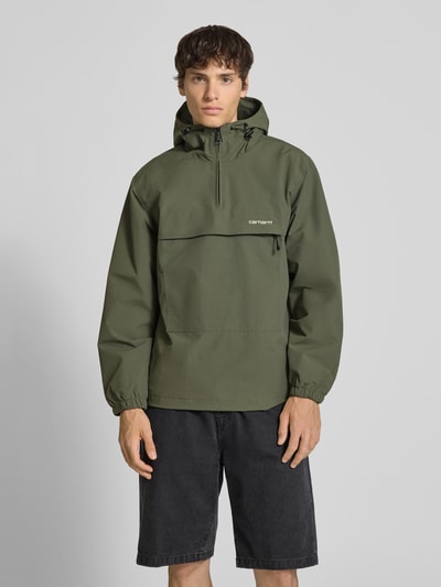 Männermodel trägt Windbreaker mit Label-Stitching von Carhartt Work In Progress in Olivgrün, Regular-Fit, mit Kapuze und Reißverschluss. Ganzkörperansicht von vorne. Kontext: Herren Windbreaker, Rückenlänge bei Größe M: 73 cm, Schulterbreite 48 cm, Brustweite 55 cm, Ärmellänge 66 cm.