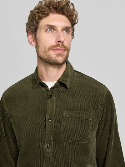 Męski model nosi Regular Fit Cordhemd mit Brusttasche od Scotch & Soda, z bawełny, kołnierzyk typu kent, brustasza, naszywka z logo, odcień oliwkowy, tekstura sztruksowa, szerokość ramion w rozmiarze M: 46 cm, długość tyłu: 78 cm, długość rękawów: 66 cm, zbliżenie na górną część ciała