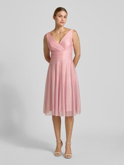 Frauenmodel trägt Knielanges Cocktailkleid mit V-Ausschnitt von TROYDEN COLLECTION in zartem Rosa, Chiffon, taillierter Schnitt, Ganzkörperansicht von vorne.
