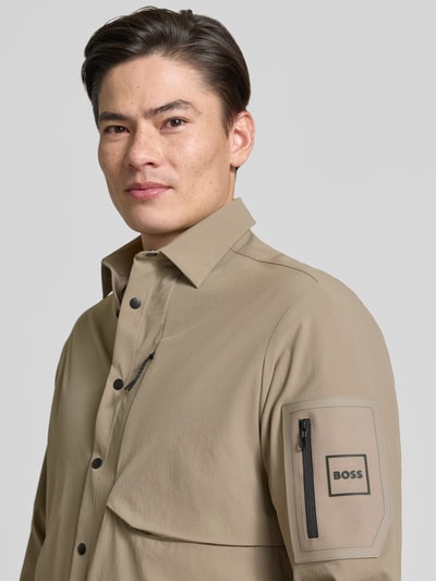 Mężczyznamodel nosi Relaxed Fit Overshirt mit Label Patch model 'ST_GLOBE HEXA L' von BOSS Green, efekt: jednolity kolor, krój relaxed fit, wykładany kołnierz, listwa z zatrzaskami, aplikacja z logo, długi rękaw, hydrofobowy, wpuszczane kieszenie boczne, brustasza, beżowy, tekstura gładka, Nahaufnahme górnej części ciała z przodu