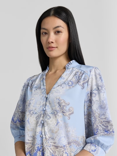 Frauenmodel trägt Damen Bluse von Neo Noir, Regular-Fit, Allover-Look, Modell 'Roselle Artist Paisley', Stehkragen, V-Ausschnitt, Knopfleiste, 3/4-Arm, zartes Blau mit Paisley, seidige Textur, Nahaufnahme Oberkörper. Rückenlänge bei Größe 36: 57 cm, Ärmellänge 43 cm.