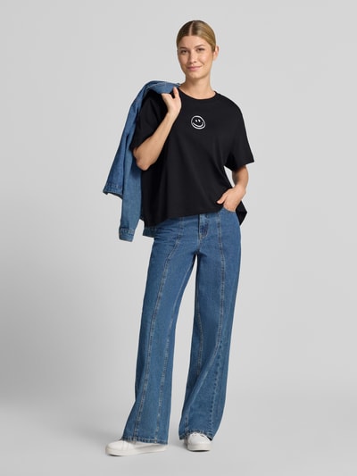 Kobiece modelka nosi czarny damski t-shirt o kroju boxy fit z czystej bawełny model 'SARA' od VERO MODA, szerokie niebieskie dżinsy, białe buty sportowe, jednolite tło, całopostaciowy widok z przodu.