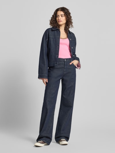 Frauenmodel trägt Slim Fit Top mit Logo-Stitching Modell 'Fiora' von OH APRIL in Pink aus elastischem Baumwoll-Mix mit Strukturmuster, kombiniert mit dunkler Jeansjacke und Hose, Rundhalsausschnitt sichtbar, Ganzkörperansicht von vorne.