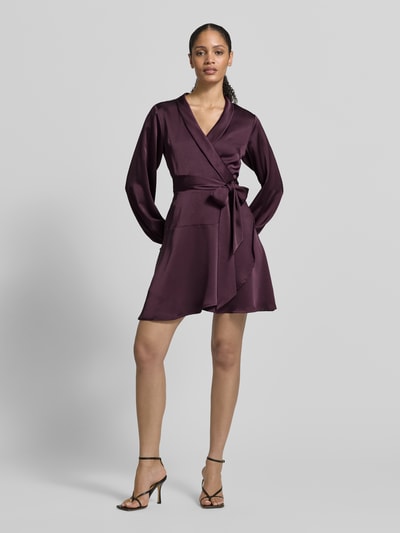 Frauenmodel trägt knielanges Kleid in Wickel-Optik Modell 'Colbie' von Neo Noir aus dunkelviolettem Satin, Tailliertes Design, Ganzkörperansicht von vorne.