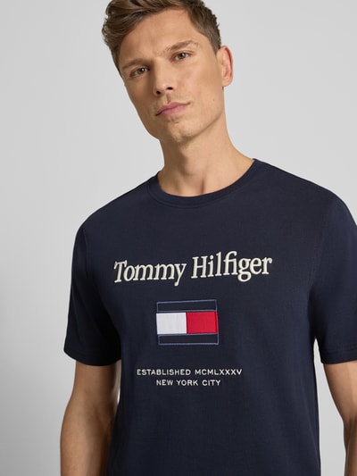 Męski model nosi Regular Fit T-Shirt aus reiner Baumwolle von Tommy Hilfiger, kolor granatowy, krótki rękaw, jersey, z przodu napis i logo, półzbliżenie górnej części ciała.