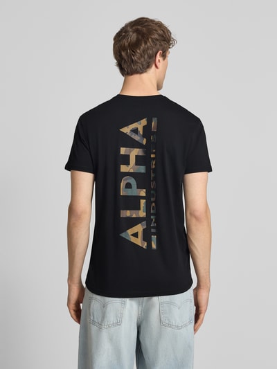 Männermodel trägt T-Shirt aus Baumwolle von Alpha Industries in Schwarz mit Label-Details, Gerader-Schnitt, Rückenlänge 67 cm, Ganzkörperansicht Rückansicht.