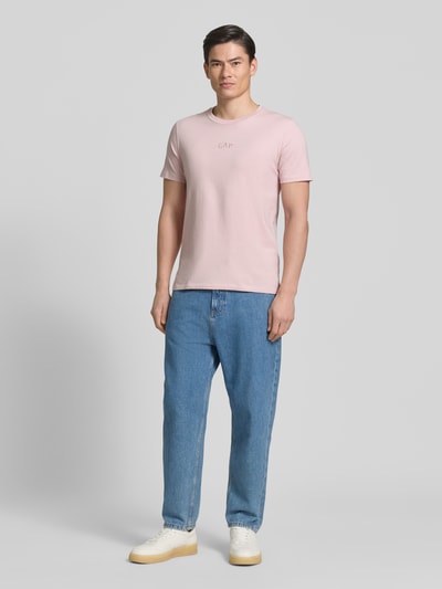 Männermodel trägt T-Shirt mit Rundhalsausschnitt und Label-Stitching von GAP in Rosa, Reine Baumwolle, Regular-Fit, Ganzkörperansicht von vorne.