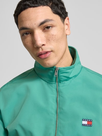 Männermodel trägt Regular Fit Blouson von Tommy Jeans in Grün mit Stehkragen, Label Patch, verdecktem Reißverschluss, Nahaufnahme Oberkörper.
