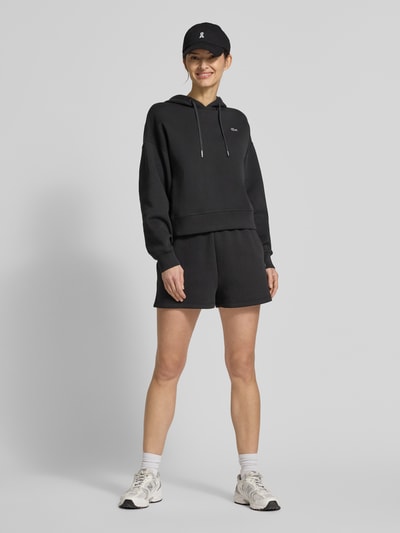 Kobiece modelka nosi Regular Fit Sweatshorts aus Baumwoll-Mix od Lacoste Sport, czarny kolor, materiał dresowy bawełniany, logo, widok całej sylwetki z przodu.