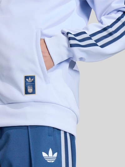 Männermodel trägt Regular Fit Trainingsjacke mit Baumwoll-Anteil Modell 'AERBLU' von adidas Originals in Optik: Unifarben mit Kontraststreifen, Logo-Stitching, Italia-Plott, Label-Patch, Stehkragen, Raglanärmel sowie Rumpfabschluss und Ärmelabschluss gerippt, Materialverarbeitung: Sweat, Eingrifftaschen, Nahaufnahme Oberkörper.