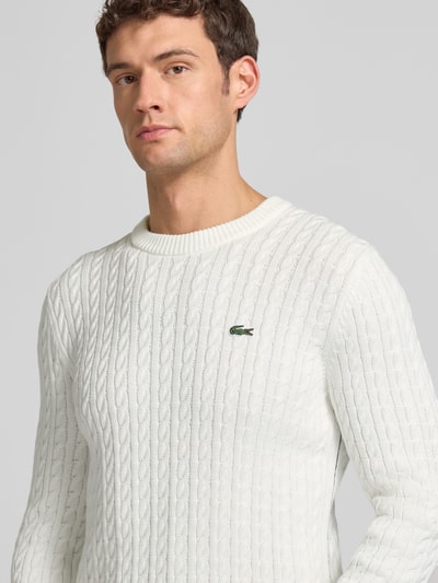 Männermodel draagt Regular Fit gebreide pullover uit katoenmix van Lacoste, effen look, kabelpatroon, logopatch, ronde hals, lange mouw, geribde boorden, detailopname bovenlichaam.