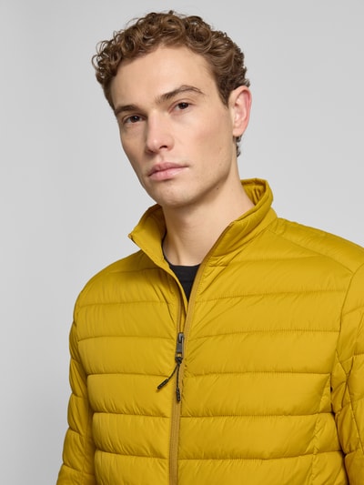Męski model nosi Steppjacke mit Reißverschluss od MCNEAL w kolorze musztardowym; tekstura pikowana, zbliżenie na górną część ciała, Regular Fit, stójka, kieszenie na zamek, długość pleców 73 cm, szerokość ramion 46 cm, szerokość klatki 57 cm, długość rękawa 65 cm.