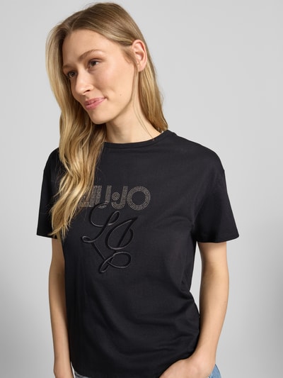 Kobiece model prezentuje T-Shirt mit Label-Stitching und Ziersteinbesatz od Liu Jo White z bawełny, krój regular fit, okrągły dekolt, długość tyłu 56 cm, zbliżenie na górną część tułowia