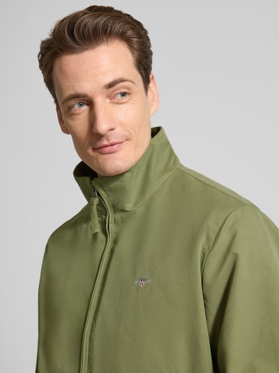 Männermodel trägt Jacke mit Stehkragen und Zweiwege-Reißverschluss von Gant, olivgrün, glatter Material-Mix, Label-Stitching, Nahaufnahme Oberkörper. Regular Fit, Baumwoll-Anteil, Eingrifftaschen, Maße Größe M: Rückenlänge 67 cm, Schulterbreite 49 cm, Brustweite 62 cm, Ärmellänge 69 cm.