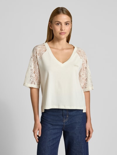 Frauenmodel trägt T-Shirt mit Spitze und Logo-Stitching von LIU JO SPORT in Creme, Raglanärmel aus Spitze, Reine Baumwolle, Gerader-Schnitt, V-Ausschnitt, verlängerter Rücken, Rückenlänge 56 cm bei Größe S, Ganzkörperansicht von vorne.