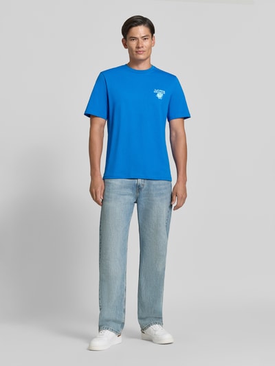 Männermodel trägt Regular Fit T-Shirt aus reiner Baumwolle Modell 'VIBING COCO' von REVIEW in Blau, Ganzkörperansicht von vorne, Herren T-Shirt von REVIEW x ANSON´S, Print auf der Rückseite, Rückenlänge bei Größe M 68 cm.