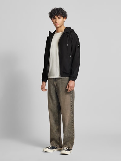 Mannenmodel draagt Softshelljacke mit Reißverschlusstaschen van Jack & Jones in zwart, regular fit, met capuchon en tunnelkoord, ritssluiting, ritszakken, ruglengte maat M 59 cm, schouderbreedte 39 cm, borstwijdte 48 cm, mouwlengte 62 cm. Ganzkörperansicht van voren.