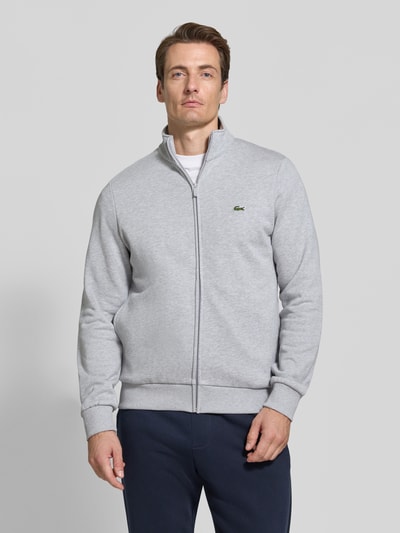 Männermodel trägt Regular Fit Sweatjacke aus Baumwoll-Mix von Lacoste in Hellgrau mit Reißverschluss, glatte Struktur, Ganzkörperansicht von vorne. Herren Sweatjacke von Lacoste.