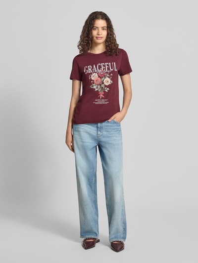 Frauenmodel trägt Regular Fit T-Shirt aus reiner Baumwolle 'ONLLUCY' von ONLY in Dunkelrot mit Blumenmuster, helle Jeans, Damen T-Shirt von ONLY, Ganzkörperansicht von vorne.